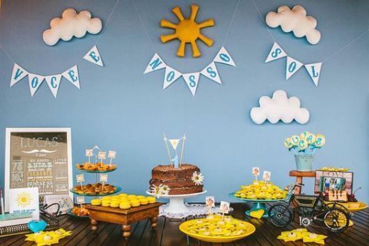 Parede azul, nuvens, sol e mesa de bolo com naked cake.