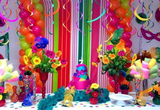 Mesa de bolo com balões, flores e serpentinas coloridas.