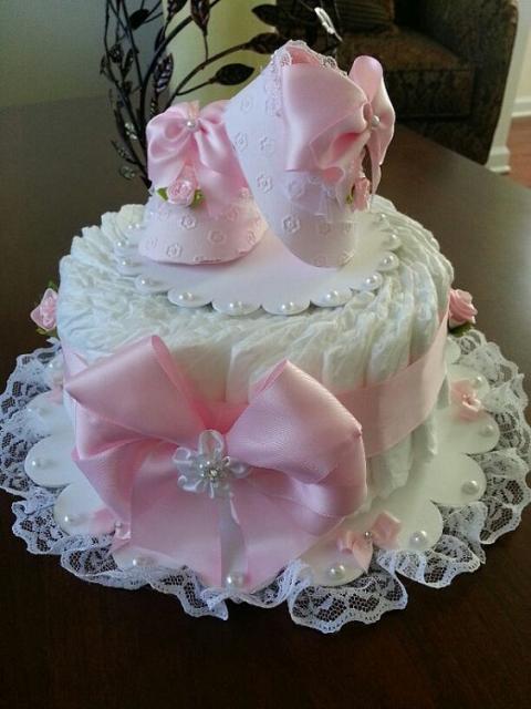 bolo fake feminino branco com um grande laço rosa e sapatinhos de bebê rosa encima
