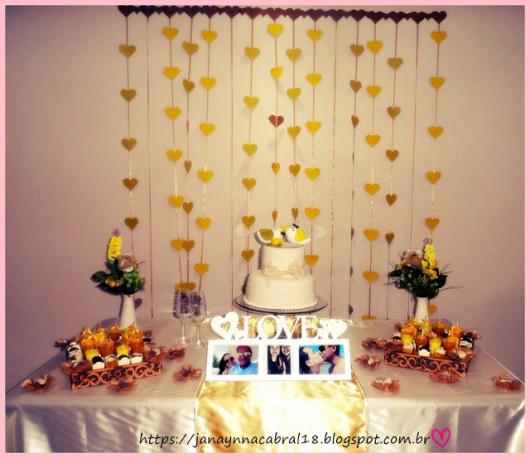 cortina de coração dourado ao fundo da mesa de doces, que conta com fotos do casal