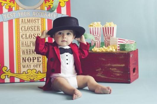 Bebê vestido de mágico, com body branco, paleto vermelho e chapéu preto.